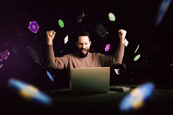 A man celebrating a winning moment on his laptop while gaming symbols float around him, capturing the excitement of BINGOMAX games.